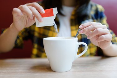 a close up of a person pouring a red and white packet of sugar into a white mug and mixing it in with a silver spoon