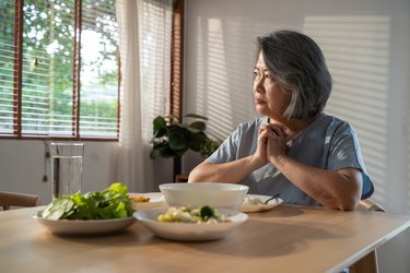 Asian retired grandmother stay at home with painful face sitting alone on eating table in house. Depressed mature Senior old woman upset feeling unhappy, lonely and missing her family at home.