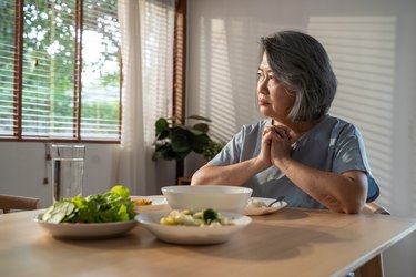 亚洲退休奶奶呆在家里，痛苦的脸独自坐在家里的餐桌上。抑郁成熟的老年妇女心烦意乱，感到不快乐，孤独和想念她的家人在家里。