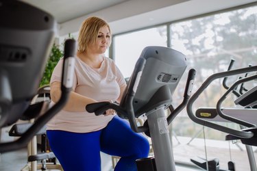 Adult exercising on bike at the gym in a white tee shirt and blue leggings