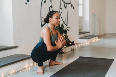 Young adult practicing yoga on a black yoga mat in garland pose (yoga squat or Malasana) in a yoga studio with twinkle lights along the baseboards