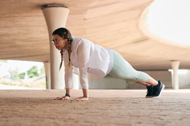 a young adult wearing light green leggings, a white long sleeve shirt and braids works the upper-body muscle groups doing push-ups outside