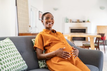 a smiling pregnant person wearing an orange jumpsuit sits on a gray couch at home holding their belly