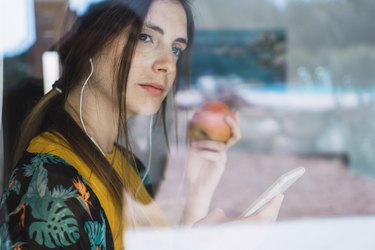 Young woman with earphones, cell phone and apple looking out of window, thinking about hunger signs