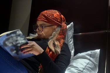 a person wearing an orange headscarf and glasses reads a book in bed propped up with several pillows to reduce acid reflux