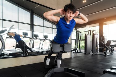 Portrait of Asian man using the glute-ham developer (GHD) while wearing blue sportswear working out on Back muscles machine at the gym. Fitness, sport, training, gym and lifestyle concept