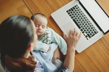 Mother holding their infant baby while typing at a computer.