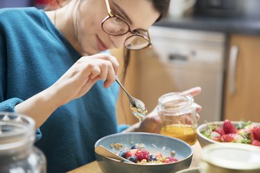 Woman pours honey over her breakfast bowl, sitting at kitchen table.