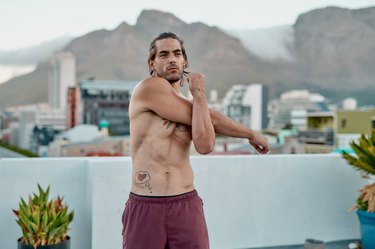 young muscular man doing dynamic arm stretches on a roof with the mountains in the background