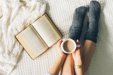 an overhead photo of a person's legs in bed wearing gray socks next to an open book with hands holding a white mug of coffee