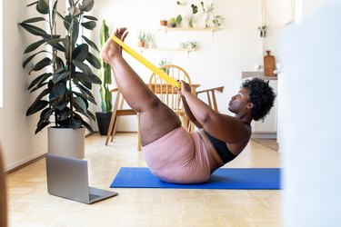 Woman doing a resistance band workout at home in her living room with computer and yoga mat