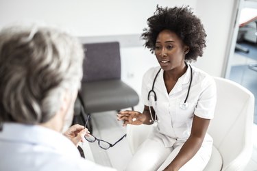 a doctor wearing white scrubs and a stethoscope talks with a patient in a waiting room with a gray chair in the background