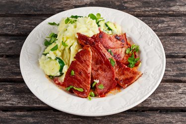 Sliced ham steak with mashed potatoes on a white plate and wooden table.