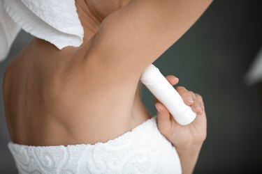 Young lady wrapped in white towel applying antiperspirant under her arms