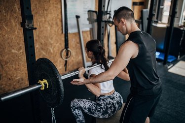 Male personal trainer assisting a woman doing a barbell back squat at the gym