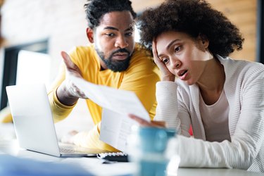 a couple looks at a laptop and paper bill at home looking stressed