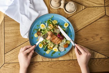 Hands with fork and knife on vegetable salad with salmon, showing eating order