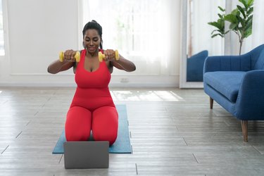 Person in a red workout outfit and ponytail doing at-home arm-toning Pilates moves with yellow hand weights