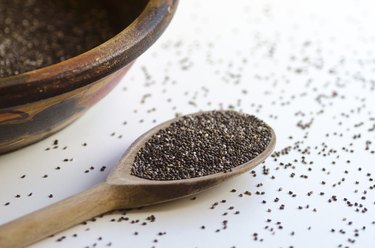 Clay vessel and wooden spoon with chia seeds on white background, seeds scattered on the table