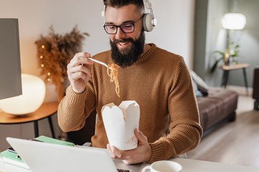 man is taking a lunch break eating noodles at desk while working from home
