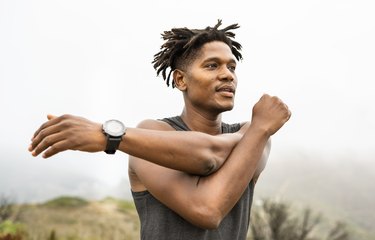 Close-up image of a person wearing a gray tank top doing a shoulder stretch outside.