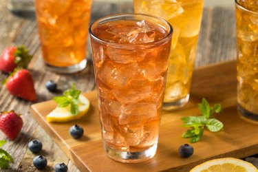 four glasses of  Iced Tea to replace water in tall clear glasses  on a wooden table and cutting board. Surrounded by strawberries, blueberries, mint leaves, and lemon slices.