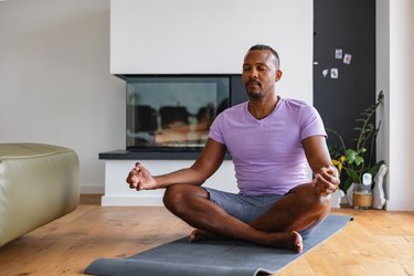 Person wearing purple T-shirt and gray shorts in easy pose on yoga mat in living room