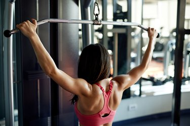Buff woman in bodybuilding performing lat pulldown exercise at machine.