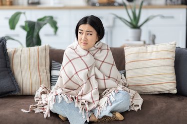 Young person wrapped in a plaid blanket sitting on the couch at home feeling cold