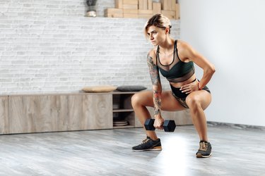 Athletic young blonde woman doing heavy dumbbell exercise in gym. Female doing squats.