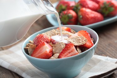 Pouring milk on top of wheat bran cereal with fresh sliced strawberries in a blue bowl