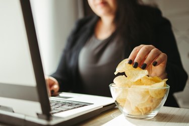 Woman's hands taking a potato chip from a bowl while she's working on a laptop