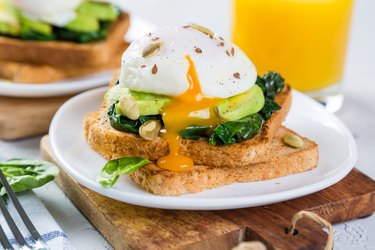 Plate of healthy breakfast including a runny poached egg on toaste with avocado and spinach on wooden cutting board.