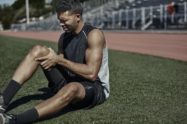 Runner wearing a black tank top and shorts sitting on grass near a track with an injured knee