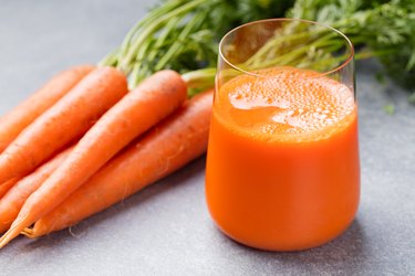 Glass of carrot juice next to raw carrots on gray countertop.
