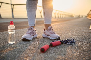 Croppped image of athlete legs wearing pink sneakers and standing on an asphalt next to a jump rope and water bottle for how to get a smaller, skinny, thinner, tiny, slim waist in 2 weeks