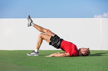 Fitness man doing body-weight glute single-leg floor bridge lift exercises. Fit athlete training glutes muscles with one-legged floor bridge butt raise in summer outdoor gym on grass