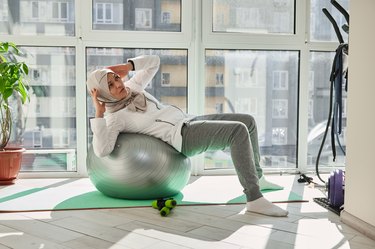 an athlete wearing a hijab doing stability ball bicycle crunches in front of windows on a green yoga mat