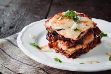 Square portion of eggplant parmesan on a white plate on wooden table.