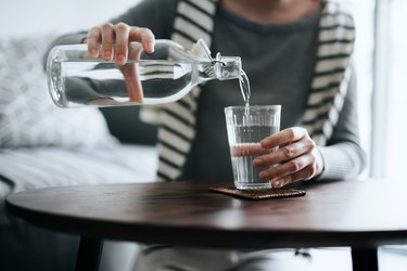 Close up of person with a gray shirt and striped sweater pouring a glass of alkaline water into a cup.