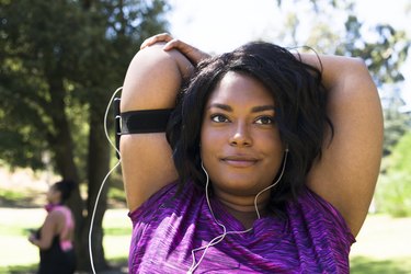 woman wearing a purple shirt and listening to headphones while doing a triceps stretch in a park