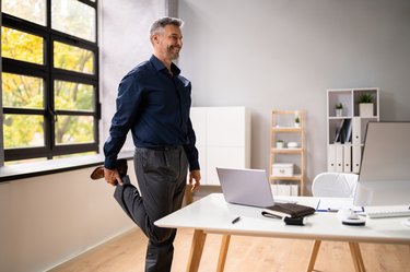 older man doing a standing quad stretch in his office