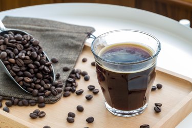 an espresso shot in a glass near a pile of coffee beans on a wooden tray on a white table
