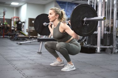 A person in an olive green tank top and leggings performing a barbell back squat in the gym