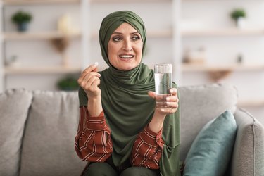 an adult wearing a hijab holding a pill and a glass of water sitting on a gray couch