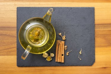 Glass pot of dalchini green tea on black platter with cinnamon sticks to illustrate green tea honey cinnamon benefits
