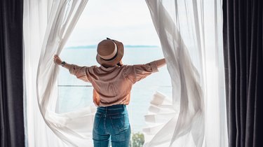 Rear view of a woman in a hotel room opening white curtains in the morning, as a way to prevent jet lag