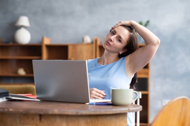 caucasian woman doing a neck and shoulder stretch in front of her laptop while working