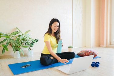 woman in yellow shirt on a blue yoga mat signing onto her laptop for an online class with a personal trainer who is trans