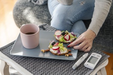 a close up photo of a person wearing jeans and a gray long sleeve shirt having breakfast of toast topped with vegetables and coffee at home on a gray tray next to a blood sugar monitor for diabetes